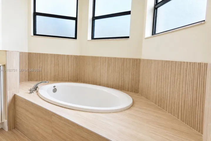 Oval soaking tub featuring vertical textured tile surround, horizontal tile decking, and metal fixtures set beneath triple windows with black frames
