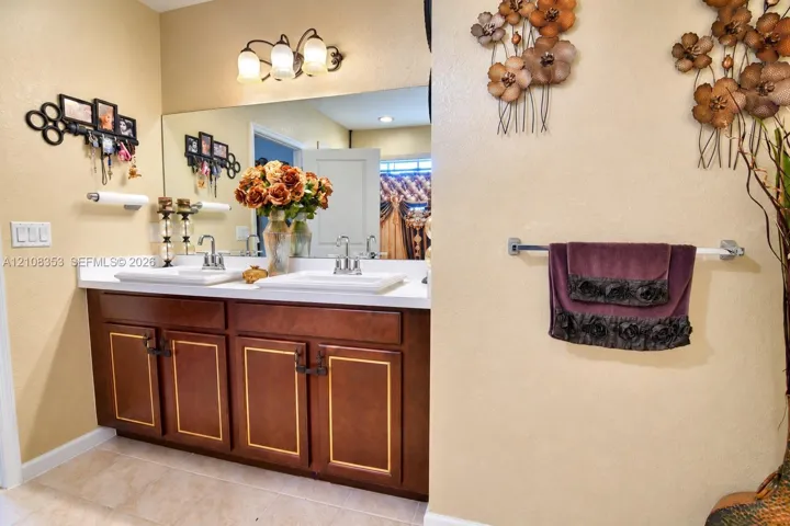 Dual-sink vanity with white surface, wood cabinetry featuring gold-toned trim, wall-mounted three-light fixture, and large rectangular mirror
