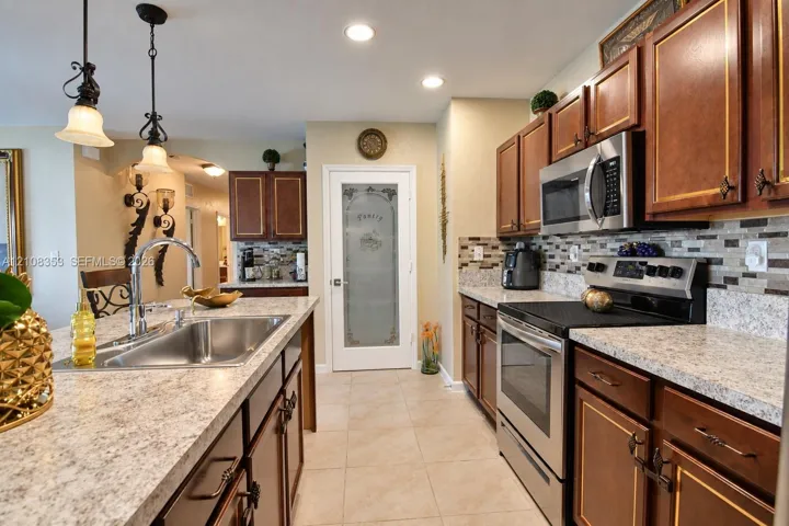 Kitchen featuring cherry-toned cabinetry, light-toned stone-finish countertops, stainless steel appliances, a multi-tone glass tile backsplash, and pantry door with frosted decorative glass