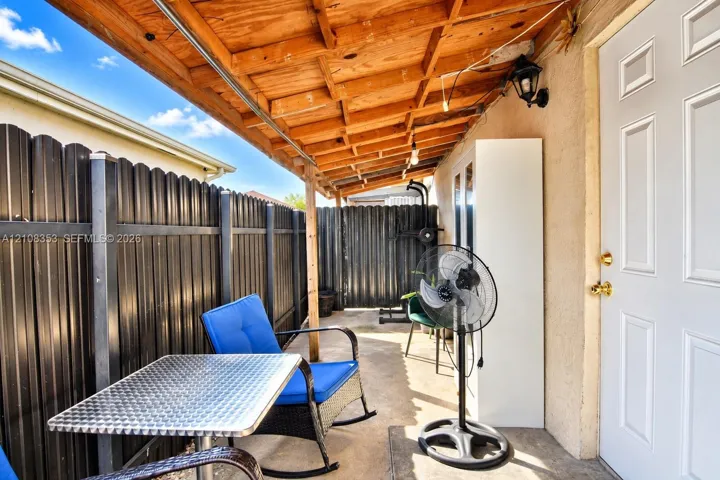 Covered concrete patio featuring a natural wood-finish rafter ceiling, black metal privacy fencing, and exterior wall-mounted lighting