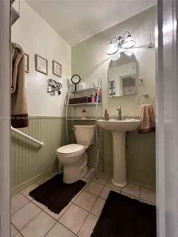 Ceramic tile flooring, pedestal sink with metal faucet, beadboard wainscoting, arched wall-mounted mirror, and three-bulb vanity lighting