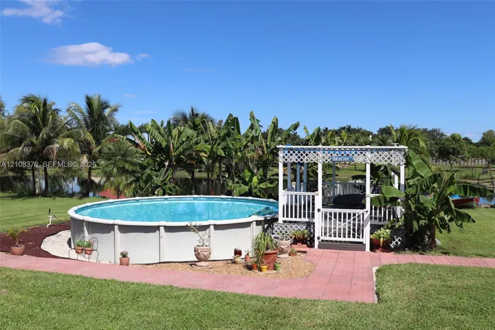Above-ground swimming pool with surrounding gravel landscaping, accompanied by a white lattice-covered gazebo featuring an integrated deck and gate