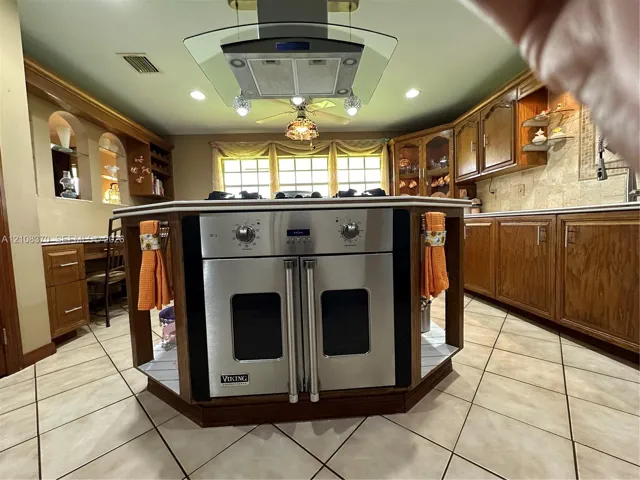 Kitchen featuring a central island with a built-in stainless steel double oven, glass-paned range hood, tile flooring, and custom cabinetry with integrated shelving