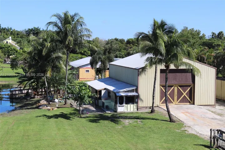 Metal-sided workshop featuring large carriage-style doors, a concrete driveway, and mature palm tree landscaping on a waterfront lot
