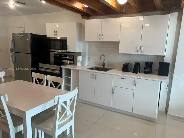 Kitchen featuring high-gloss white cabinetry, light-toned solid surface countertops, a white subway tile backsplash, stainless steel appliances, and exposed wood ceiling beams