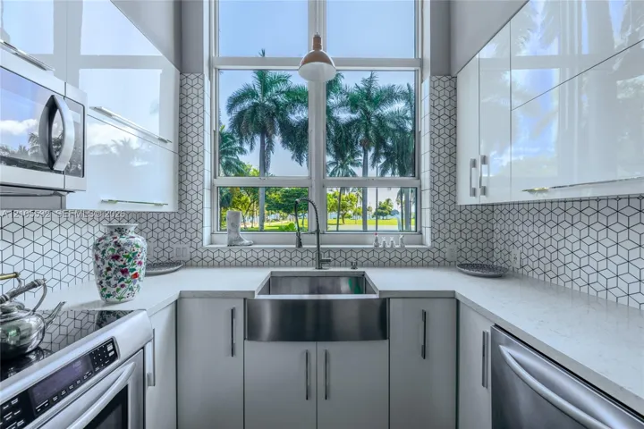 Kitchen featuring high-gloss cabinetry, stone-finish countertops, stainless steel farmhouse sink, and geometric-patterned backsplash tile, accented by a floor-to-ceiling window
