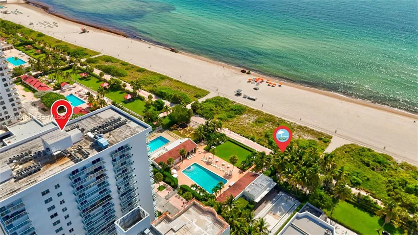 Aerial perspective of a high-rise building situated on a direct beachfront property, featuring an outdoor swimming pool, paved sundeck, and landscaped lawn areas