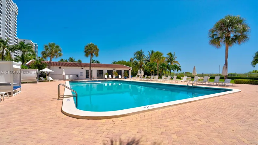 In-ground swimming pool surrounded by a paver deck, palm trees, and a low-profile clubhouse with a clay tile roof