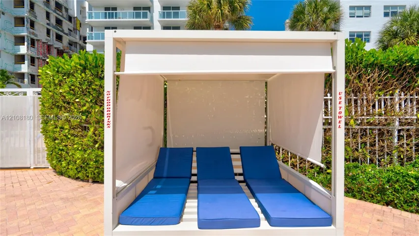 Permanent poolside cabana featuring a white metal frame, integrated retractable fabric shades, and tiered cushioned platforms situated on a brick paver deck