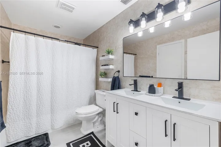 Dual-sink vanity with white cabinetry, matte black hardware, stone-surface countertop, and rectangular undermount sinks, complemented by a large-format mirror with integrated multi-bulb vanity lighting and neutral tile wall cladding