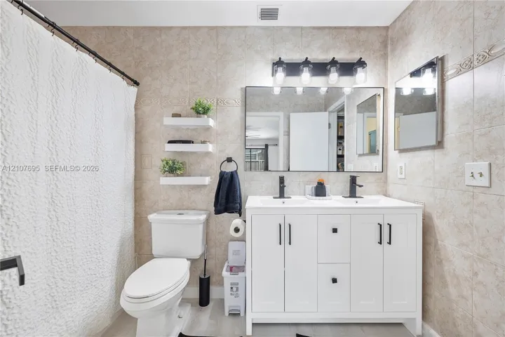 Double-sink vanity with white cabinetry, matte black hardware, and rectangular undermount sinks, complemented by a wide vanity mirror, four-light wall fixture, and neutral-toned wall tiling