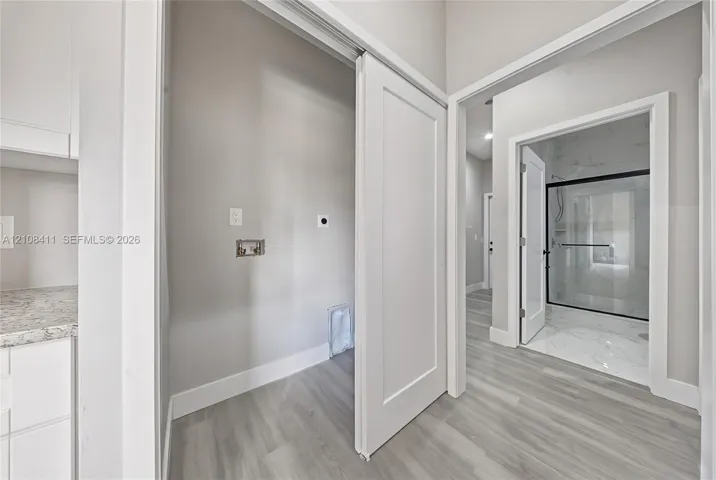 Utility space featuring light-toned wood-finish flooring, white cabinetry with speckled stone-finish countertops, and a sliding door, with a glimpse of a walk-in shower containing light-toned stone-finish tile and a black-framed glass enclosure