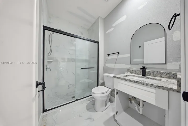 Bathroom featuring a walk-in shower with glass enclosure, stone-finish vanity countertop, arched wall mirror, and matte black hardware