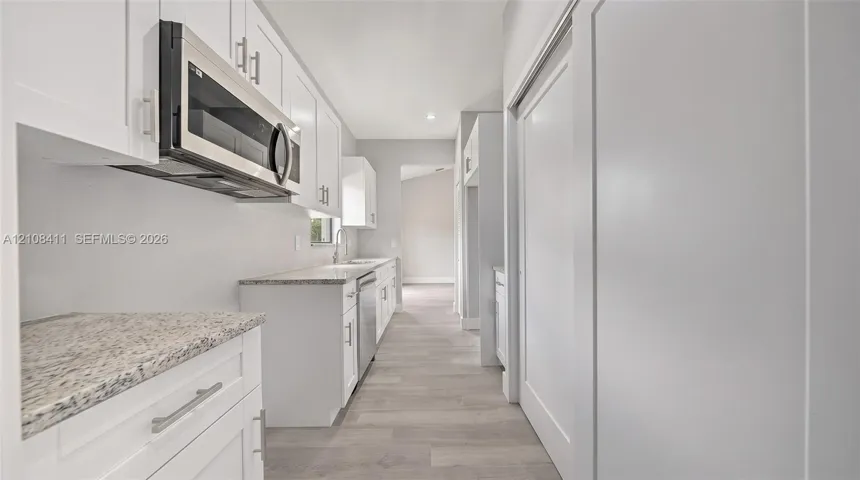 White shaker-style cabinetry featuring satin nickel hardware, speckled stone-finish countertops, stainless steel over-the-range microwave, built-in dishwasher, and wood-finish flooring