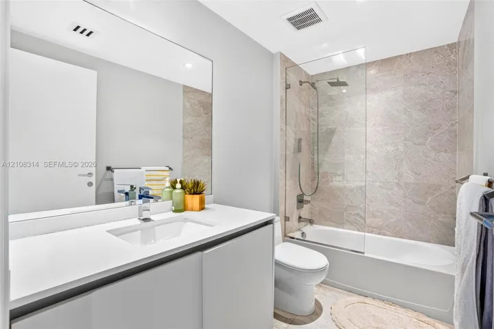 Bathroom featuring a floating vanity with a stone countertop, an integrated rectangular sink, a frameless glass shower panel, and stone-tile tub surround