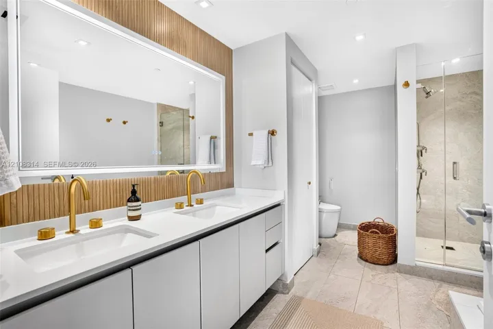 Dual-sink vanity with white surface, metallic gold-finish faucets, large-scale vanity mirror with integrated lighting, wood-slat wall paneling, and walk-in shower with glass enclosure