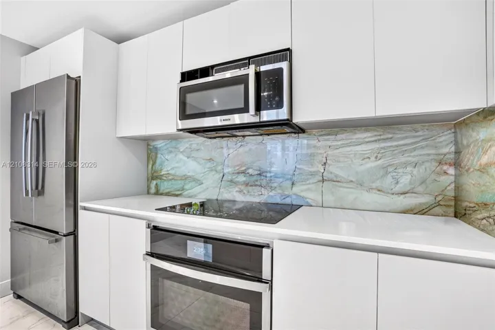 Kitchen featuring flat-panel white cabinetry, stone-patterned slab backsplash, quartz countertops, and stainless steel appliances including a built-in oven, electric cooktop, and over-the-range microwave