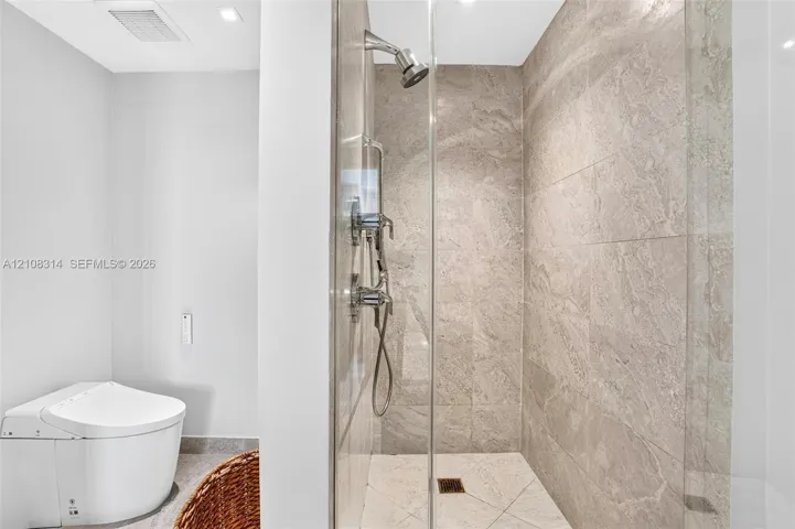Walk-in shower with floor-to-ceiling stone-finish tile, glass enclosure, and wall-mounted shower system, paired with a one-piece low-profile toilet