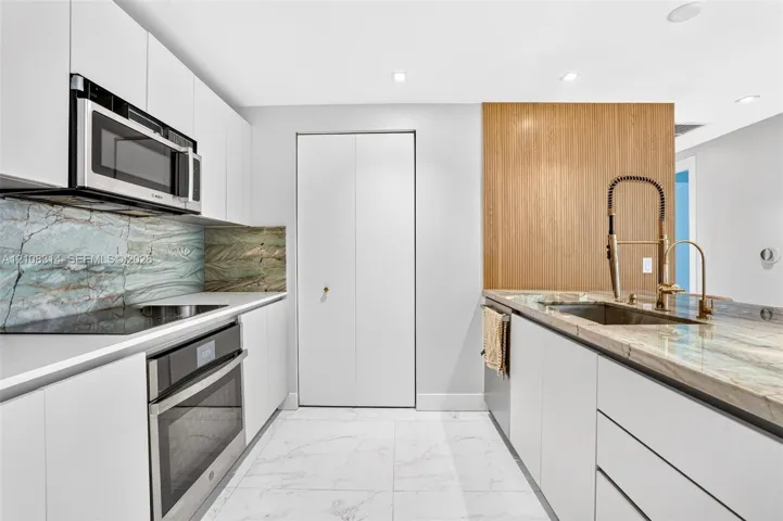 Modern kitchen featuring flat-panel white cabinetry, stone slab countertops, integrated stainless steel appliances, and wood-finish accent paneling with a gooseneck faucet