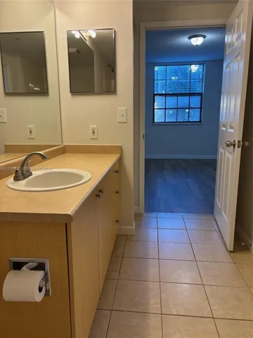 Bathroom featuring a laminate countertop, integrated sink with a curved faucet, light wood-finish cabinetry, ceramic tile flooring, and a flat-panel mirror