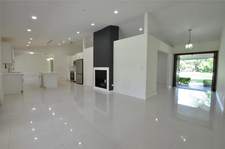 Open-concept interior featuring large-format polished tile flooring, vaulted ceilings with recessed lighting, a modern floor-to-ceiling fireplace with high-contrast surround, and stainless steel kitchen appliances