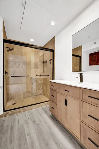 Walk-in shower with stone tile surround, glass enclosure, and matte black fixtures, paired with a wood-finish vanity, solid surface countertop, and recessed lighting