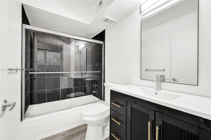 Bathroom featuring a modern vanity with a solid-surface countertop, an integrated rectangular sink, black cabinetry with gold-tone hardware, a large frameless mirror, and a shower-tub combination with dark tile surround