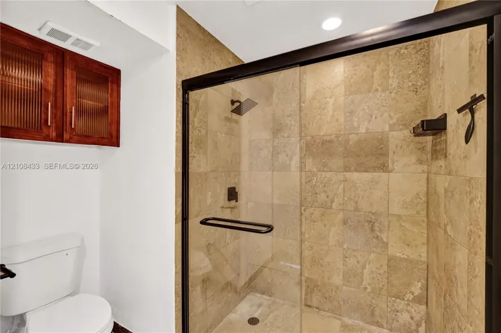 Walk-in shower with natural stone tiling, matte black frame, glass door, and overhead rainfall showerhead, complemented by wall-mounted wood-finish cabinetry