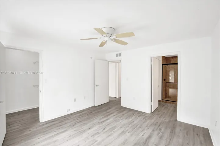 Neutral interior featuring wood-finish flooring, a ceiling fan with integrated lighting, and multiple interior doorways leading to a walk-in closet and a bathroom with a glass-enclosed shower