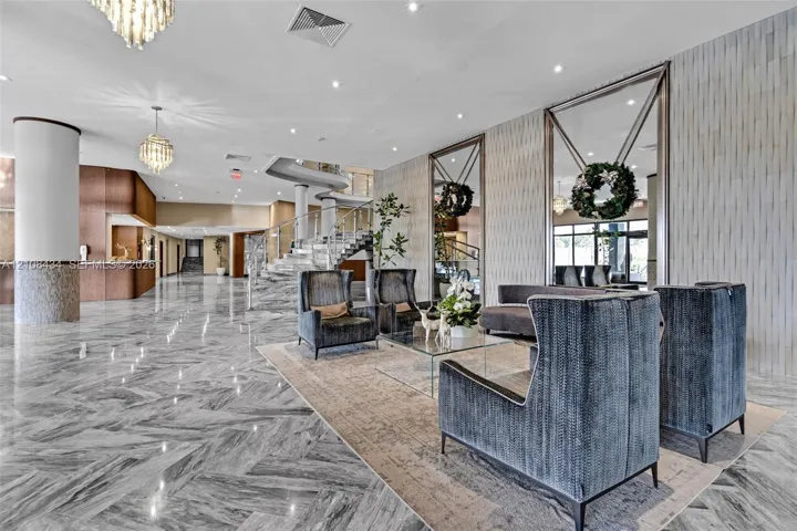 Expansive lobby featuring polished stone flooring in a herringbone pattern, floor-to-ceiling decorative mirrors, recessed lighting, crystal chandeliers, and a sweeping staircase with a metal railing
