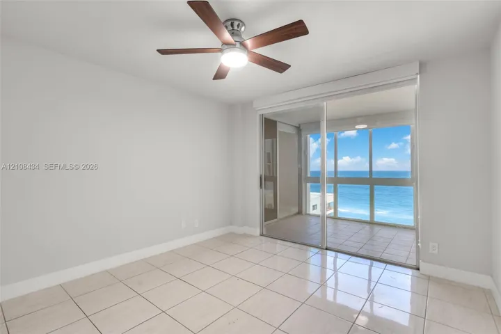 Interior space featuring large-format ceramic tile flooring, ceiling fan with integrated lighting, and full-height sliding glass doors opening to a tiled exterior balcony with an ocean backdrop