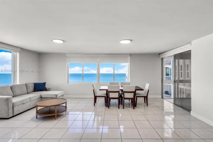 Open-concept room featuring light-toned tile flooring, large multi-pane windows, and a sliding glass door providing access to a balcony with an ocean backdrop