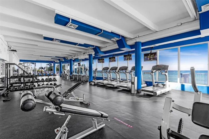 Fitness center featuring floor-to-ceiling windows with ocean scenery, exposed white beam ceilings, industrial-style exposed ductwork, and resilient rubber flooring