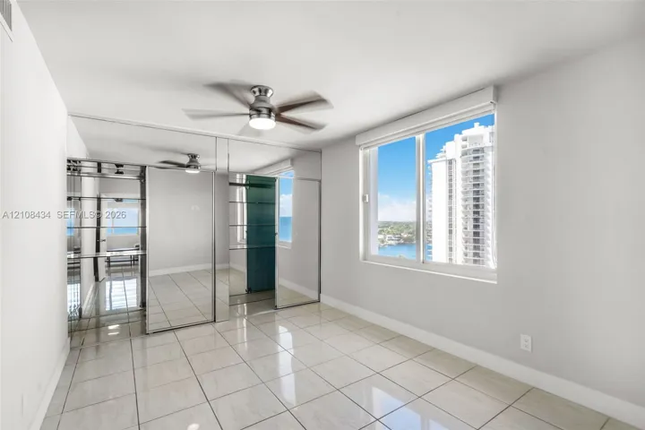 Large format tile flooring, floor-to-ceiling mirrored closet doors, ceiling fan with integrated lighting, and expansive window with water and skyline exposure