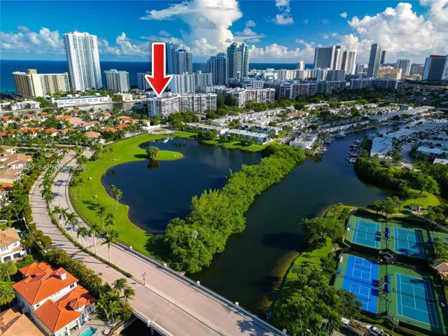Aerial perspective of a multi-story residential building featuring proximity to a central lake, landscaped grounds, tennis courts, and coastal waterway