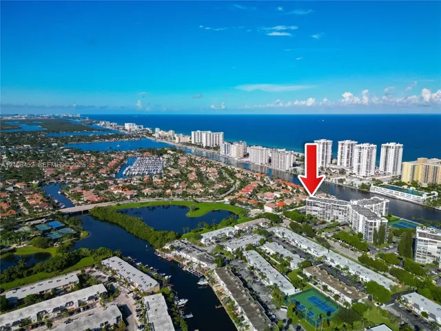 Aerial perspective of a multi-story residential building featuring waterfront positioning, proximity to coastal shoreline, boat slips, and tennis courts