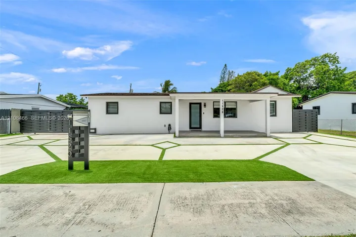 Single-story exterior featuring white stucco siding, a covered front porch, geometric concrete driveway with synthetic grass inlays, and dual metal privacy fencing