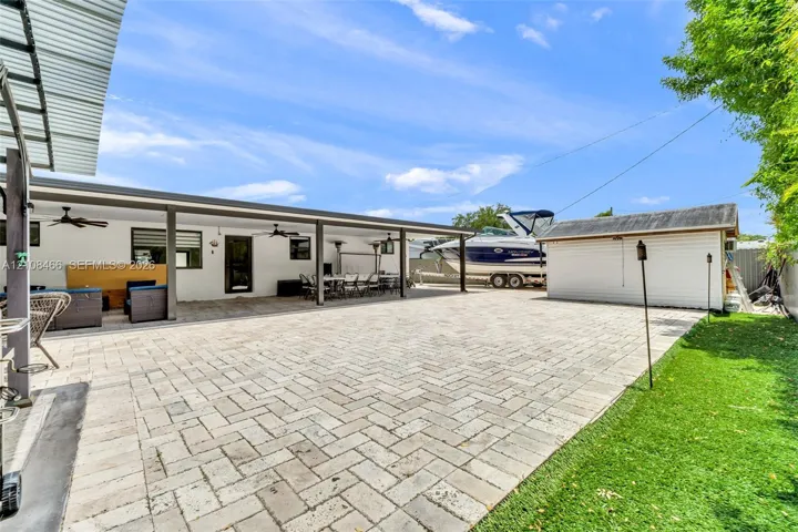 Expansive paver patio featuring a covered outdoor living area with ceiling fans, a standalone storage structure, and manicured green space
