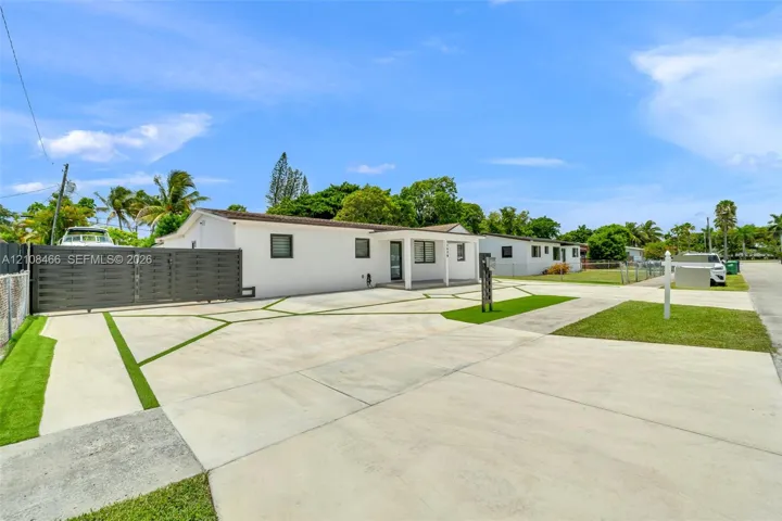 Single-story residence featuring white stucco exterior, expansive concrete driveway with geometric turf accents, horizontal slat privacy fencing, and covered front entryway