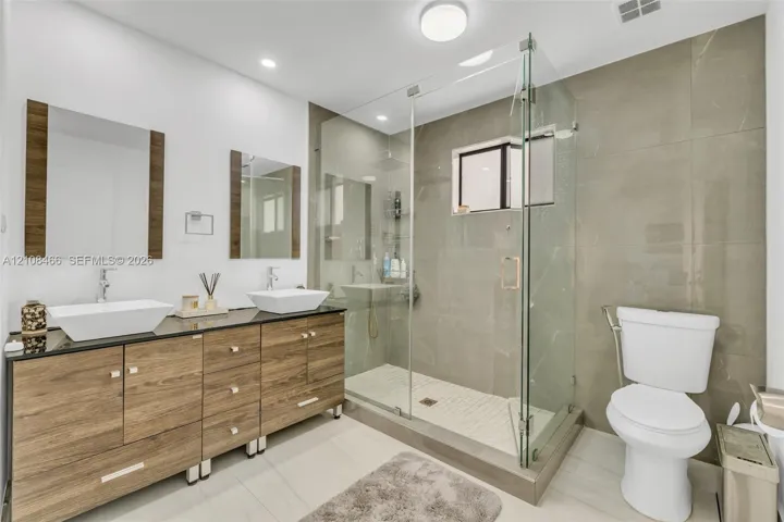 Dual vanity with rectangular vessel sinks, dark stone countertop, wood-finish cabinetry, walk-in glass-enclosed shower featuring full-height tile surround, and recessed lighting