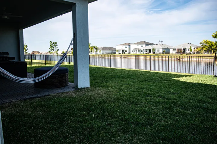 Covered patio with paver flooring, manicured lawn, and aluminum perimeter fencing bordering a waterfront