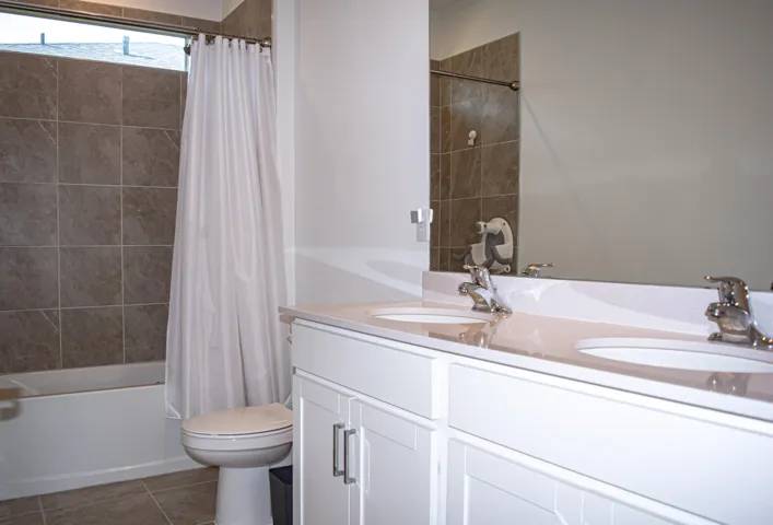 Double-sink vanity with a solid-surface countertop, white cabinetry with silver hardware, full-width wall mirror, and tile-surround shower-tub combination