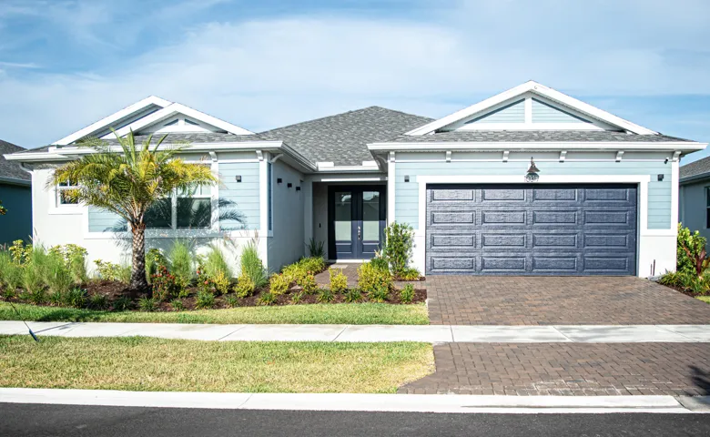 Single-story exterior featuring blue horizontal lap siding, a two-car paneled garage door, a paver driveway, double front entry doors, and gable rooflines