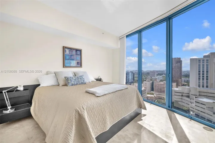 Floor-to-ceiling windows featuring urban scenery, expansive light-toned tile flooring, and a sleek, built-in dark finish headboard
