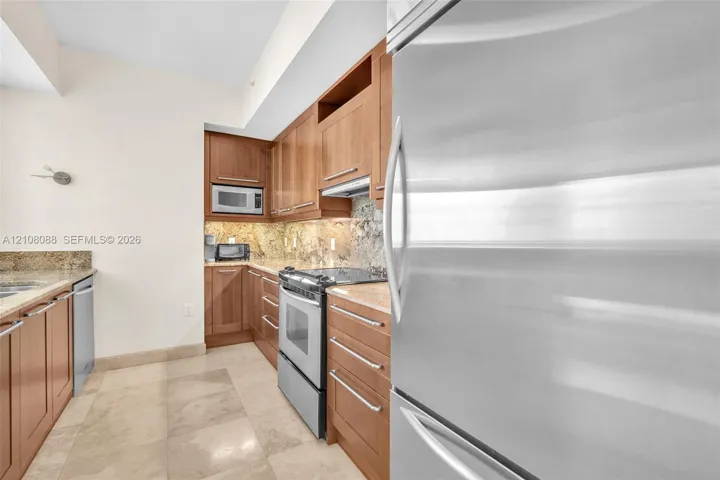 Kitchen featuring natural wood cabinetry, stone countertops, stone tile backsplash, stainless steel appliances, and large-format tile flooring