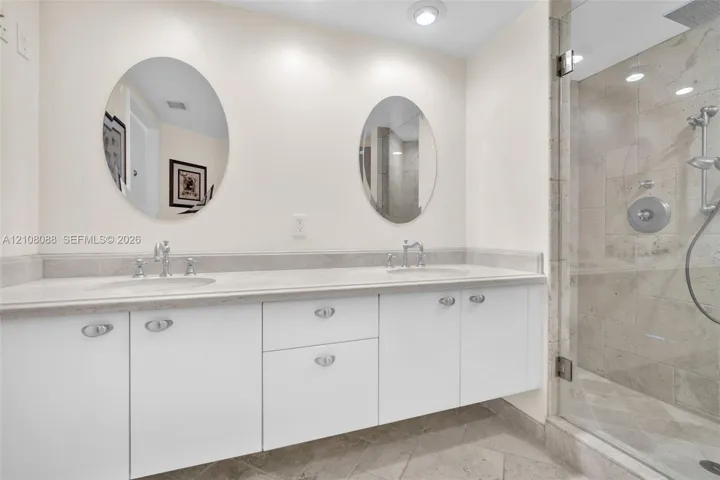 Dual-sink vanity with white cabinetry, light-toned stone-finish countertop, dual oval mirrors, and a glass-enclosed shower featuring stone wall tiles