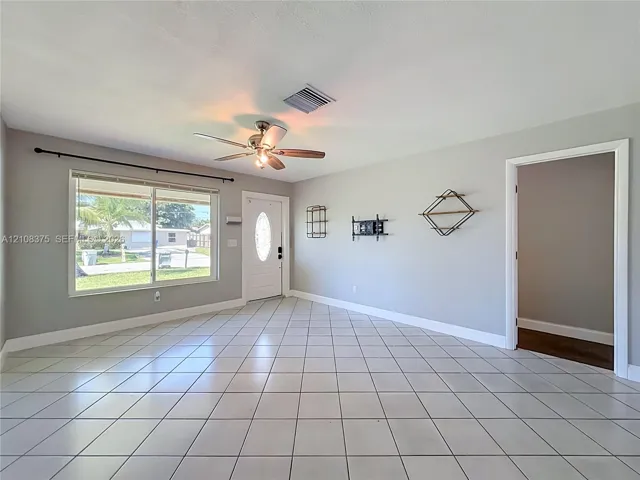 Neutral-toned interior featuring large-format ceramic tile flooring, a multi-pane window with hardware, a solid-panel front door with an oval glass insert, and a ceiling-mounted fan with integrated lighting