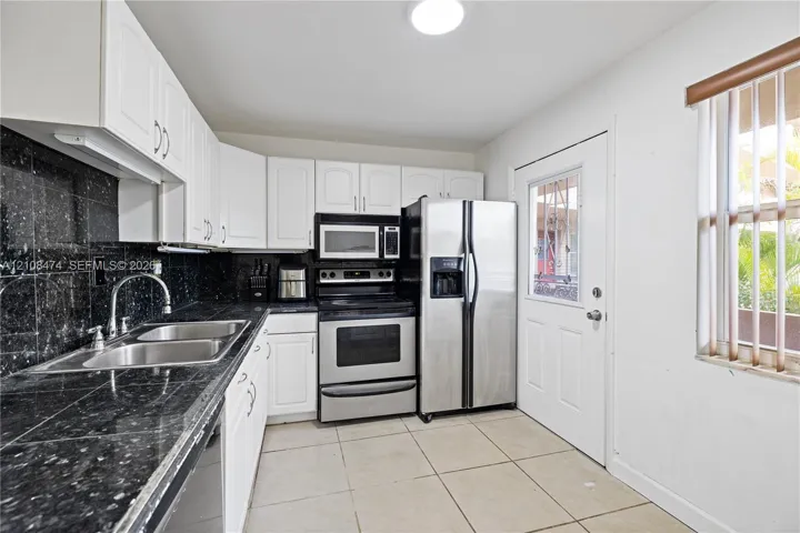 Kitchen featuring white shaker-style cabinetry, dark stone-finish countertops, tiled backsplash, stainless steel appliances, and large-format floor tile