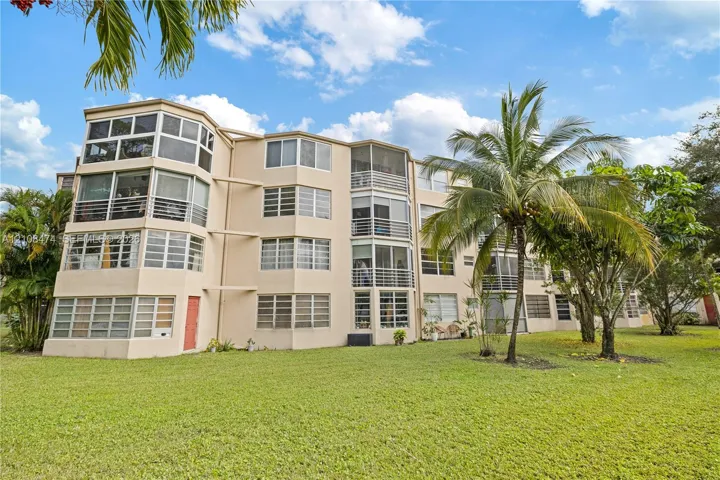 Mid-rise exterior featuring multi-level screened-in lanais, light-toned stucco facade, and expansive lawn landscaping with mature palm trees