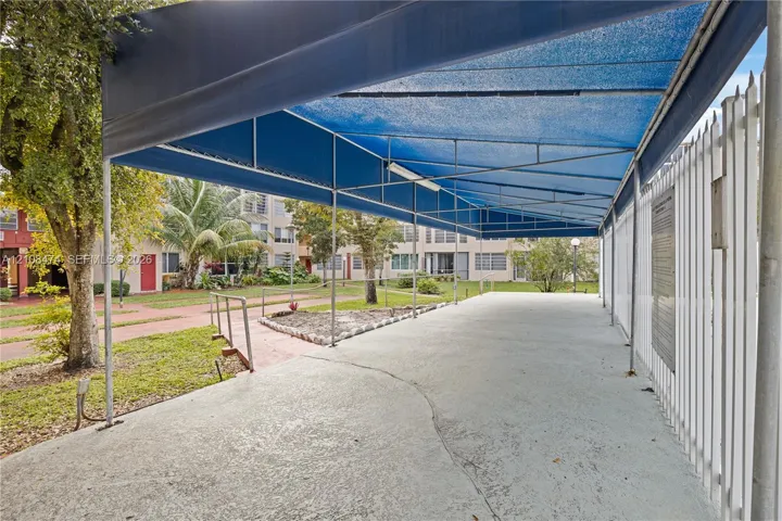 Covered outdoor patio featuring a metal frame structure, blue shade canopy, concrete flooring, and perimeter metal fencing